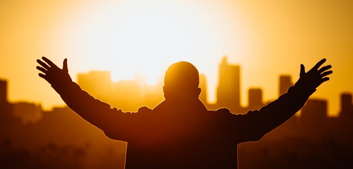 Man extending arms to sky in front of sunset over city