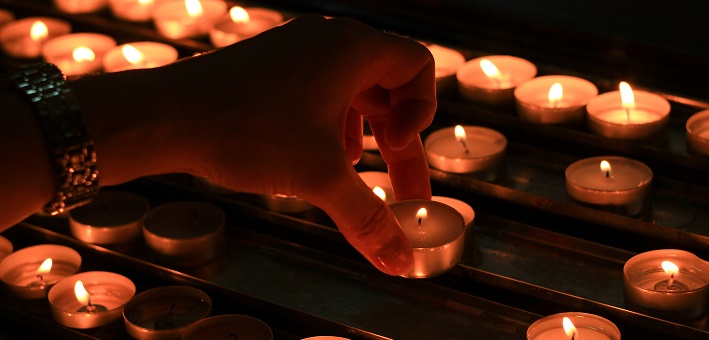 Hand placing lit votive candle among bank of candles at vigil.