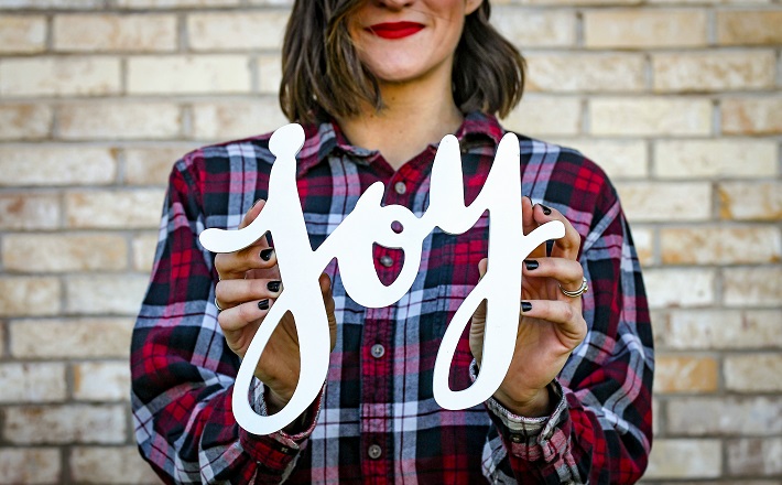 Smiling woman holding Joy cutout in front of brick wall