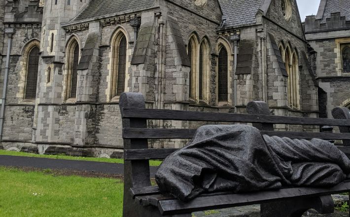 Bronze sculpture of homeless person sleeping on bench in front of church