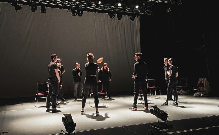 Actors dressed in black standing on starkly lit stage rehearsing