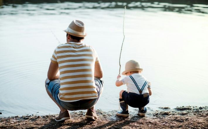 Man and young boy squatting by side of lake with fishing poles